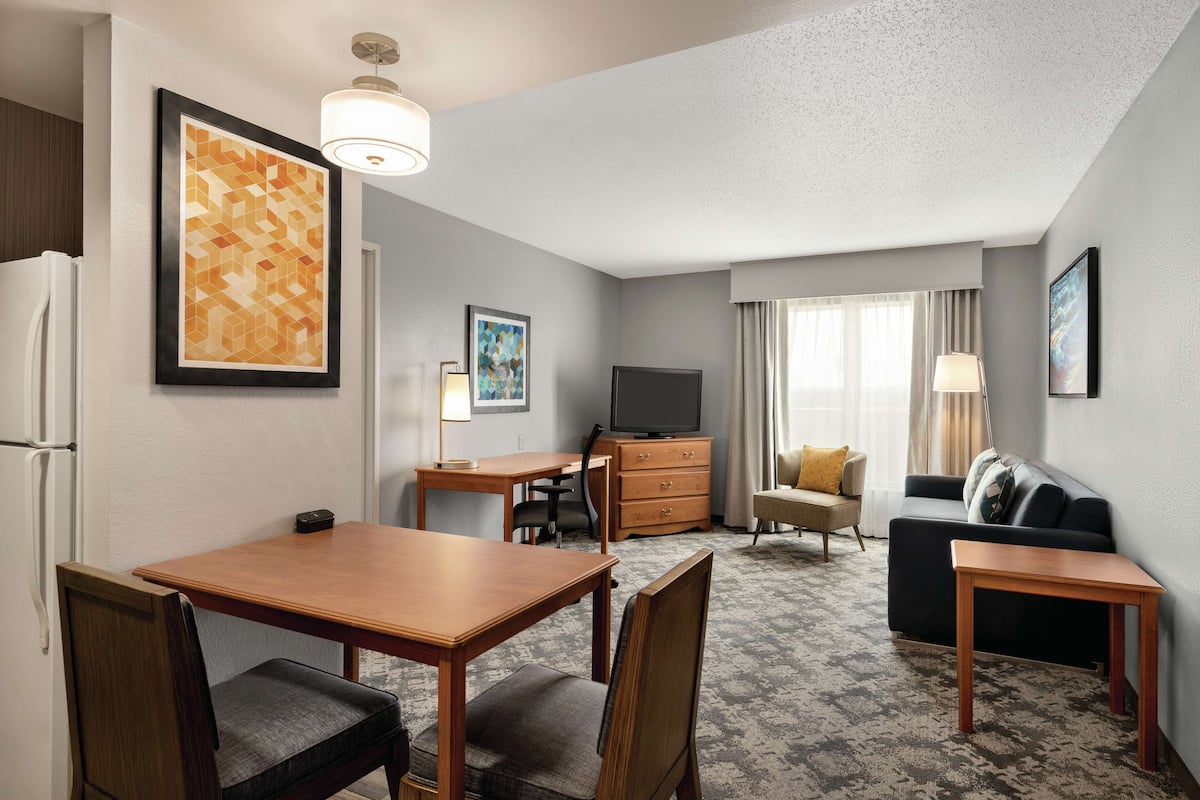 A hotel room with a dining table, chairs, a sofa, a TV, a lamp, and artwork on the walls.