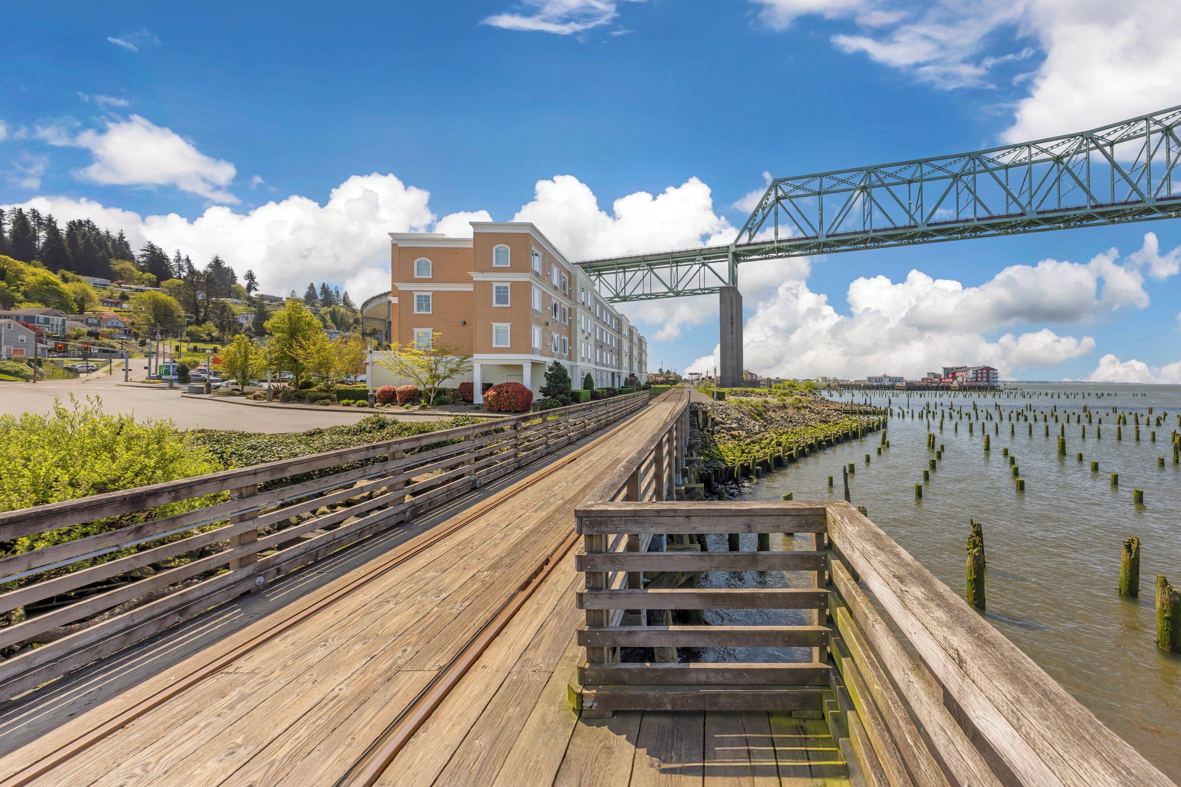 A wooden boardwalk leading to a bridge over water, with buildings and greenery in the background.