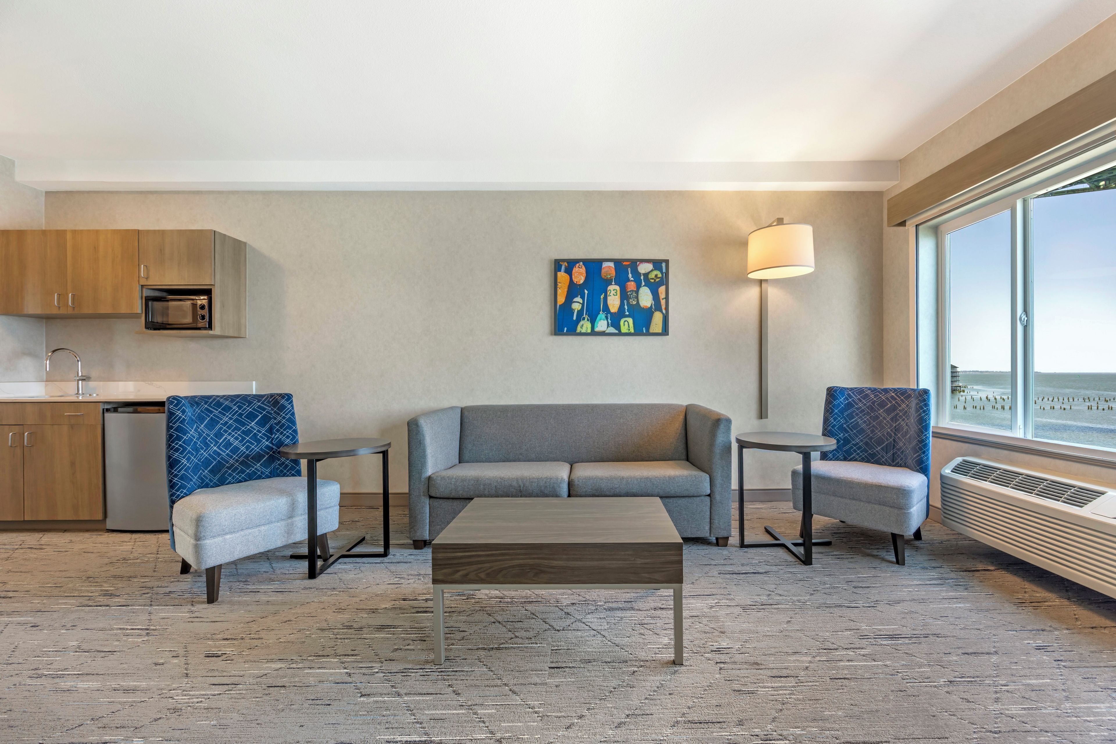 A modern hotel room with a sofa, two armchairs, a coffee table, a microwave, a small dining table, a floor lamp, and a large window.