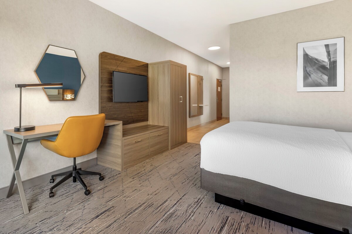 A modern hotel room with a wooden desk, an orange office chair, a bed, a TV, and a framed picture on the wall.