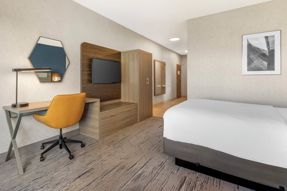 A modern hotel room with a wooden desk, an orange office chair, a bed, a TV, and a framed picture on the wall.