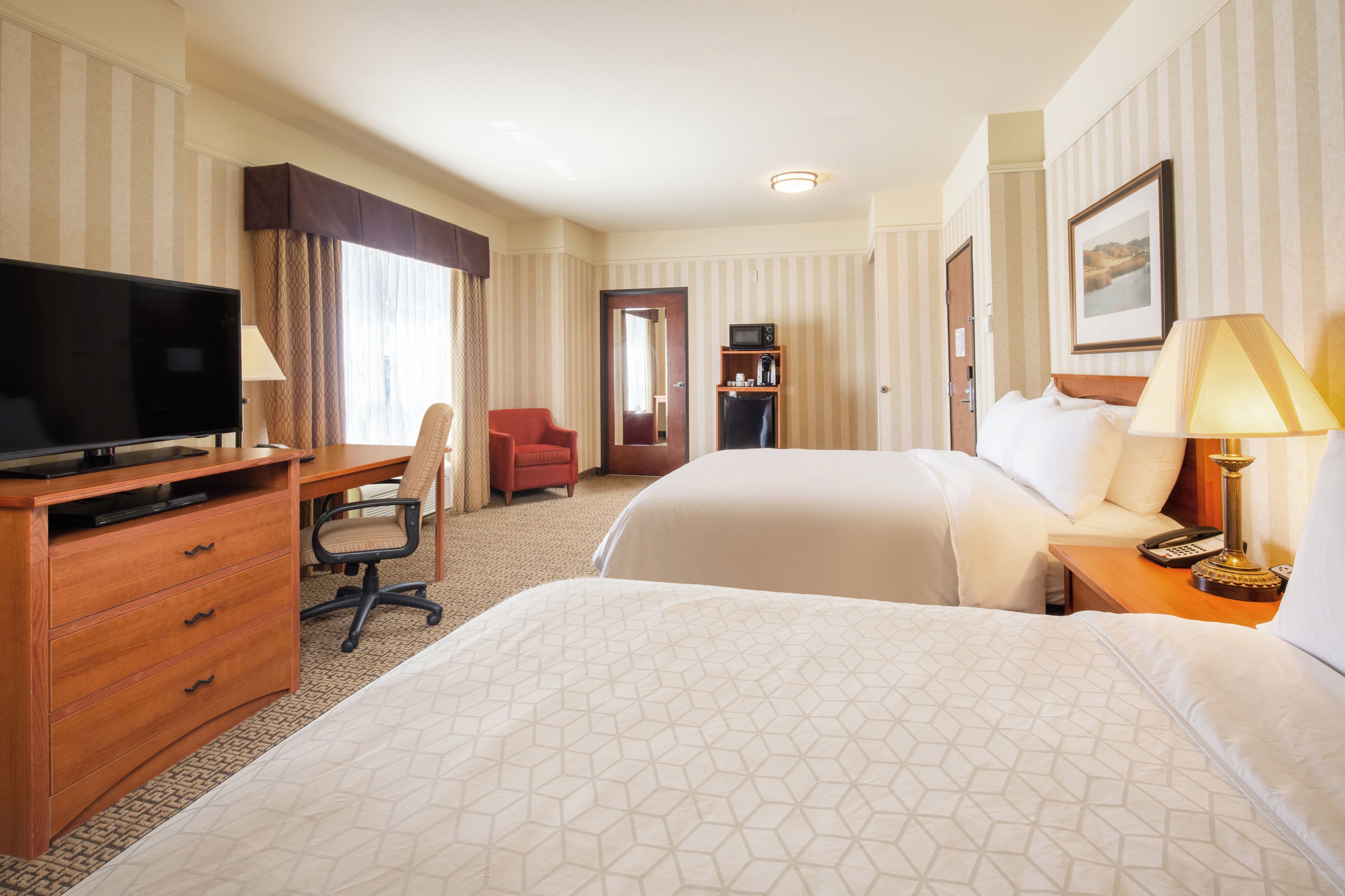 A hotel room with a bed, a desk with a television, a chair, a wardrobe, and a bedside table with a lamp.