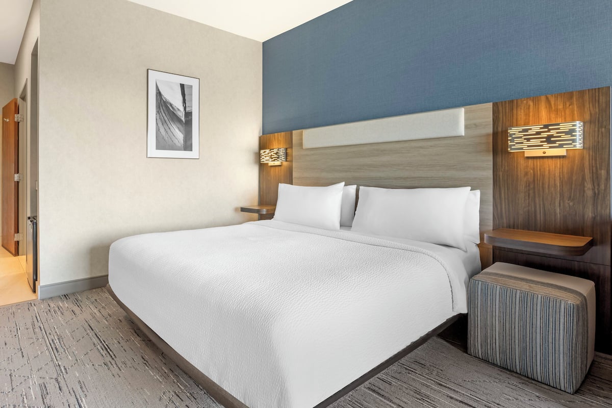 A modern hotel room with a large bed, a headboard with built-in lighting, a small bedside table, and a framed picture on the wall.