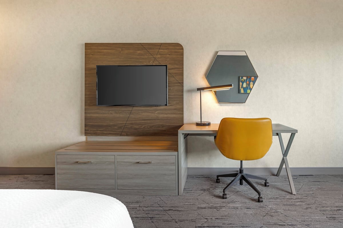 A hotel room with a bed, a desk with a chair, a flat-screen TV, a desk lamp, and a wall-mounted artwork.