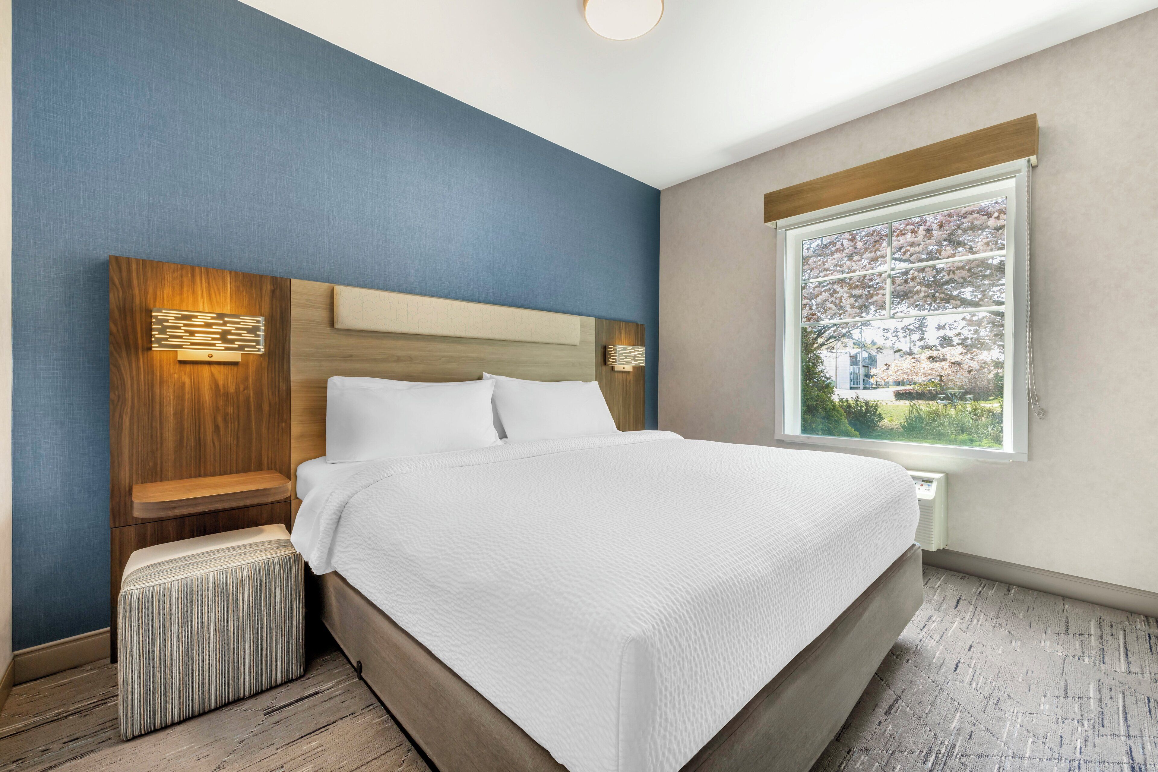 A modern hotel room with a large bed, a wooden headboard, a nightstand, and a window with a view of trees.