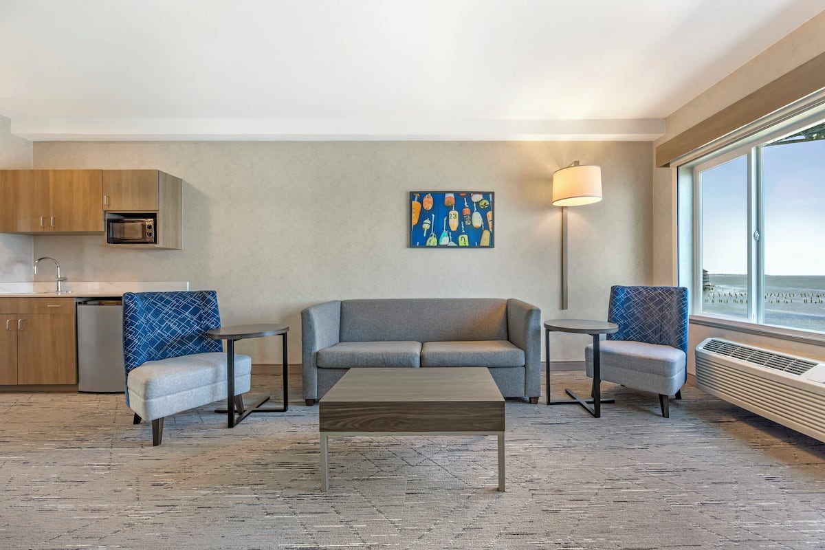 A modern hotel room with a sofa, two armchairs, a coffee table, a microwave, a small dining table, a floor lamp, and a large window.