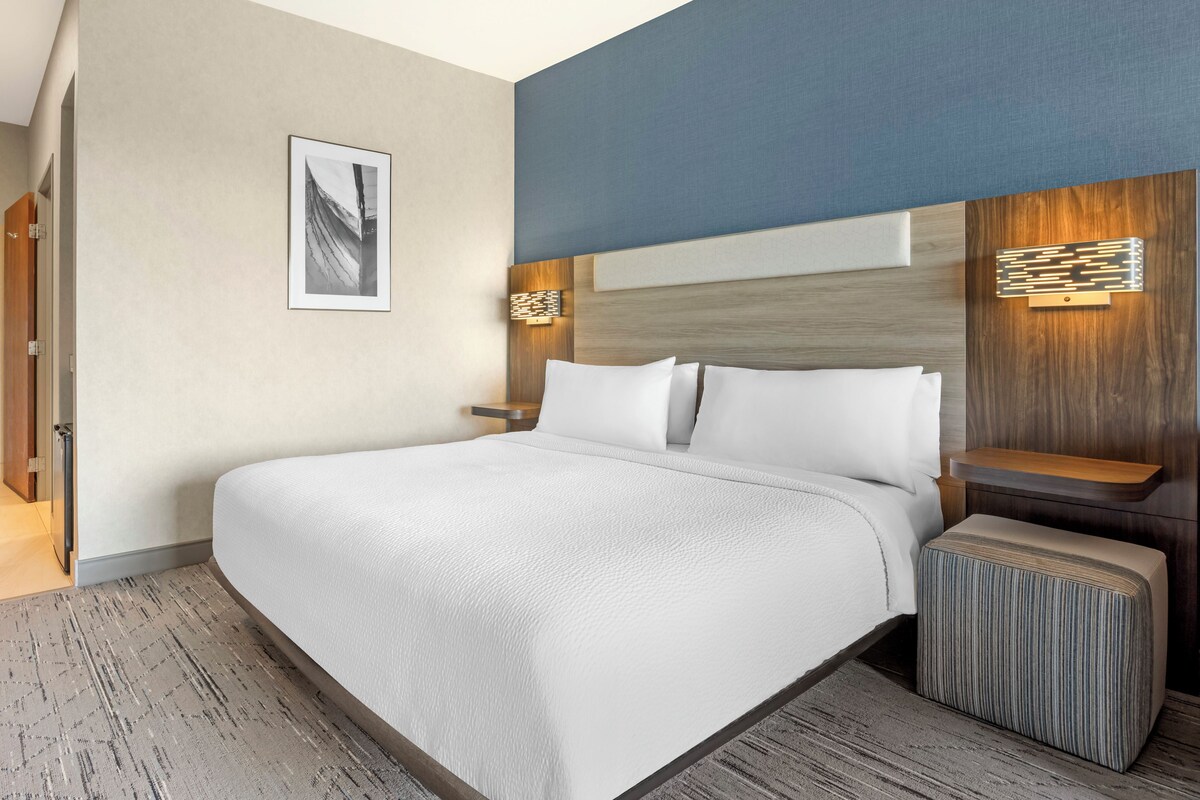 A modern hotel room with a large bed, a headboard with built-in lighting, a small bedside table, and a framed picture on the wall.