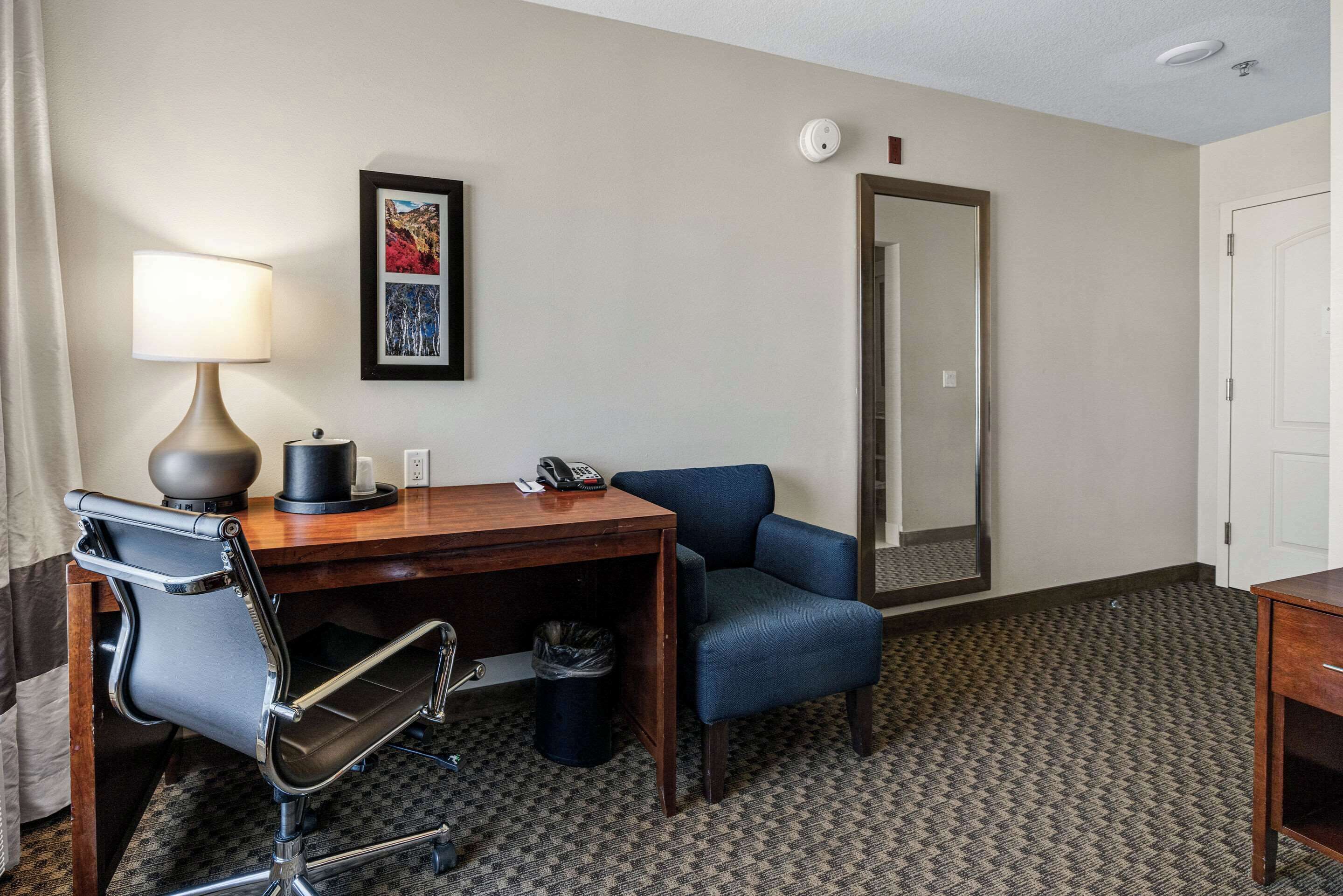 A hotel room with a wooden desk, a blue chair, a lamp, a telephone, a trash bin, a framed picture, and a mirror.