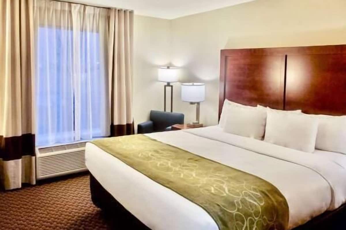 Ein Hotelzimmer mit einem großen Bett, einem Schreibtisch, einem Stuhl, einer Lampe und einem Fenster mit Vorhängen.