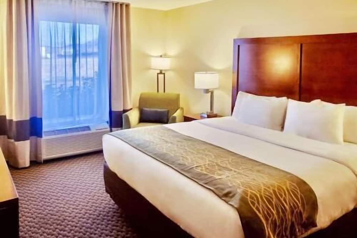 Ein Hotelzimmer mit einem großen Bett, einem Sessel, einem Schreibtisch, einer Lampe und einem Fenster mit Vorhängen.