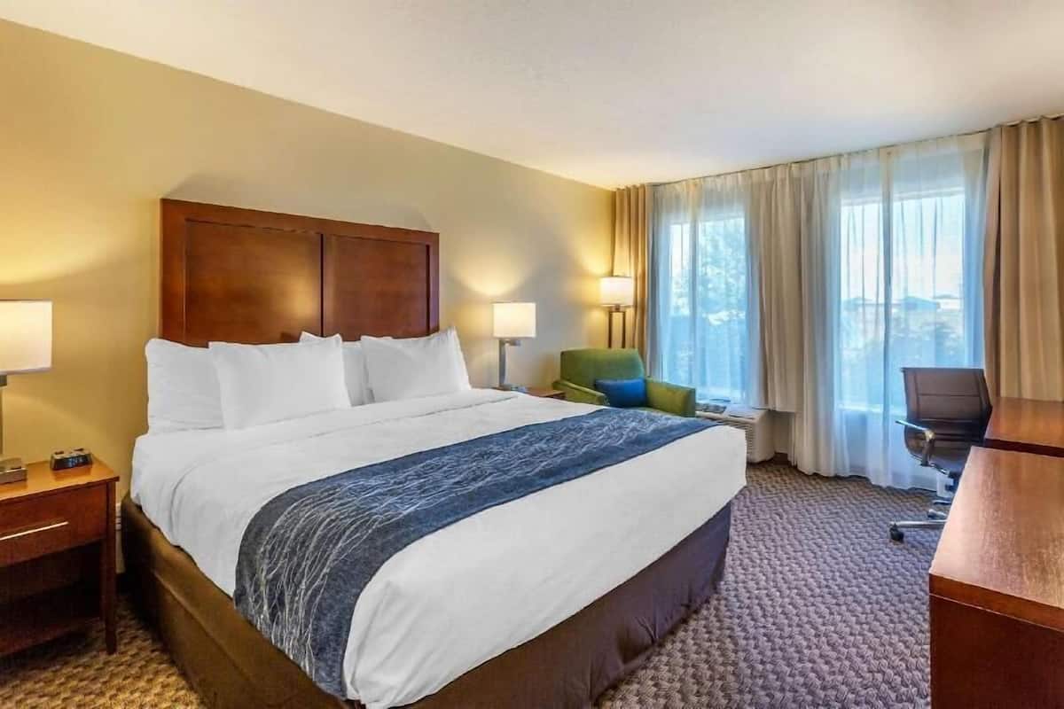 Ein Hotelzimmer mit einem großen Bett, zwei Nachttischlampen, einem Schreibtisch, einem Sessel und Blick ins Freie durch das Fenster.