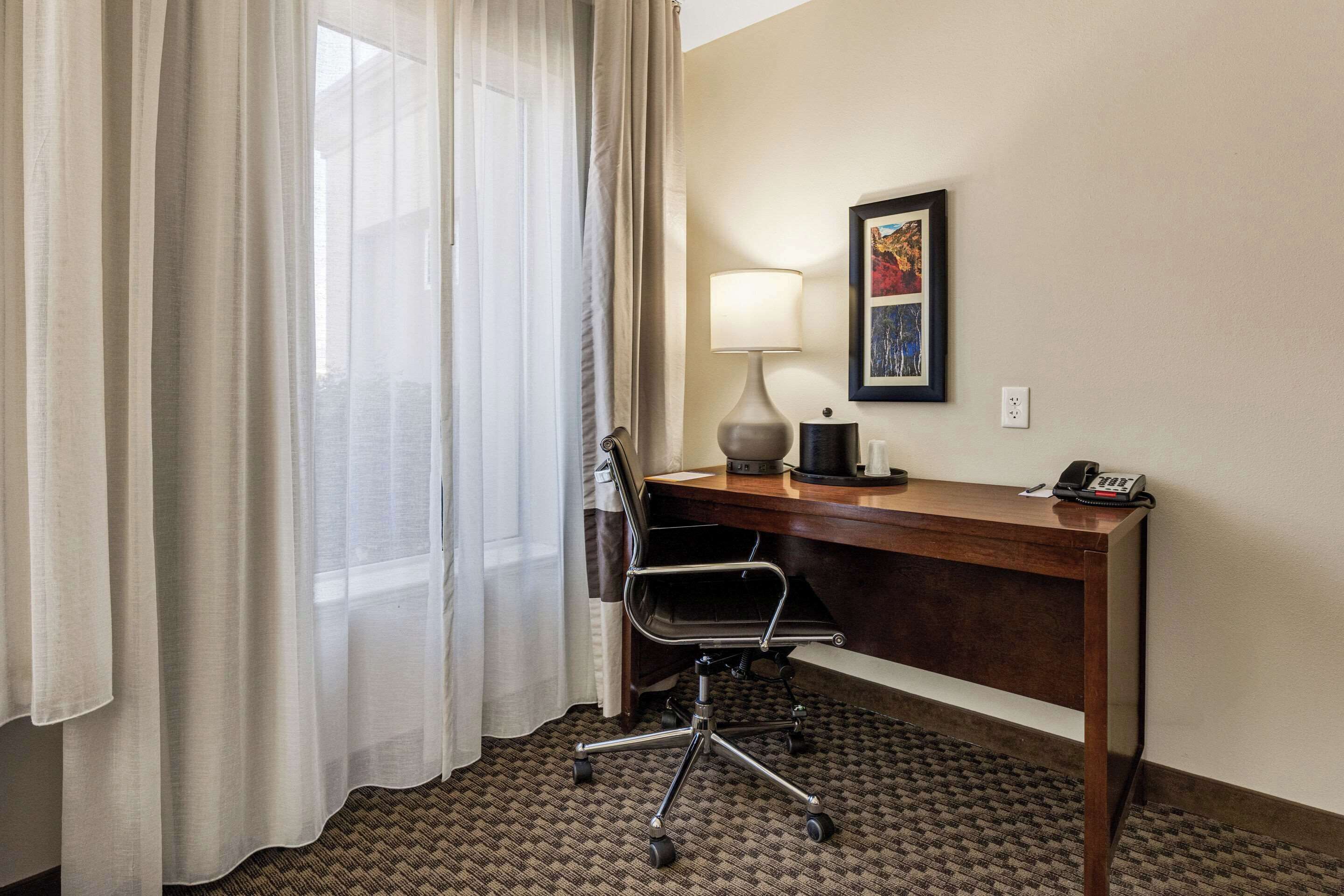 Quarto de hotel com escrivaninha, cadeira, luminária, telefone e um quadro na parede.