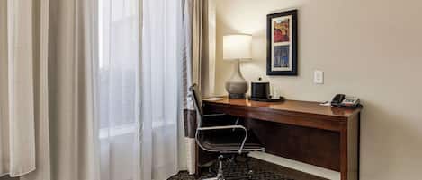 A hotel room with a desk, chair, lamp, phone, and a framed picture on the wall.