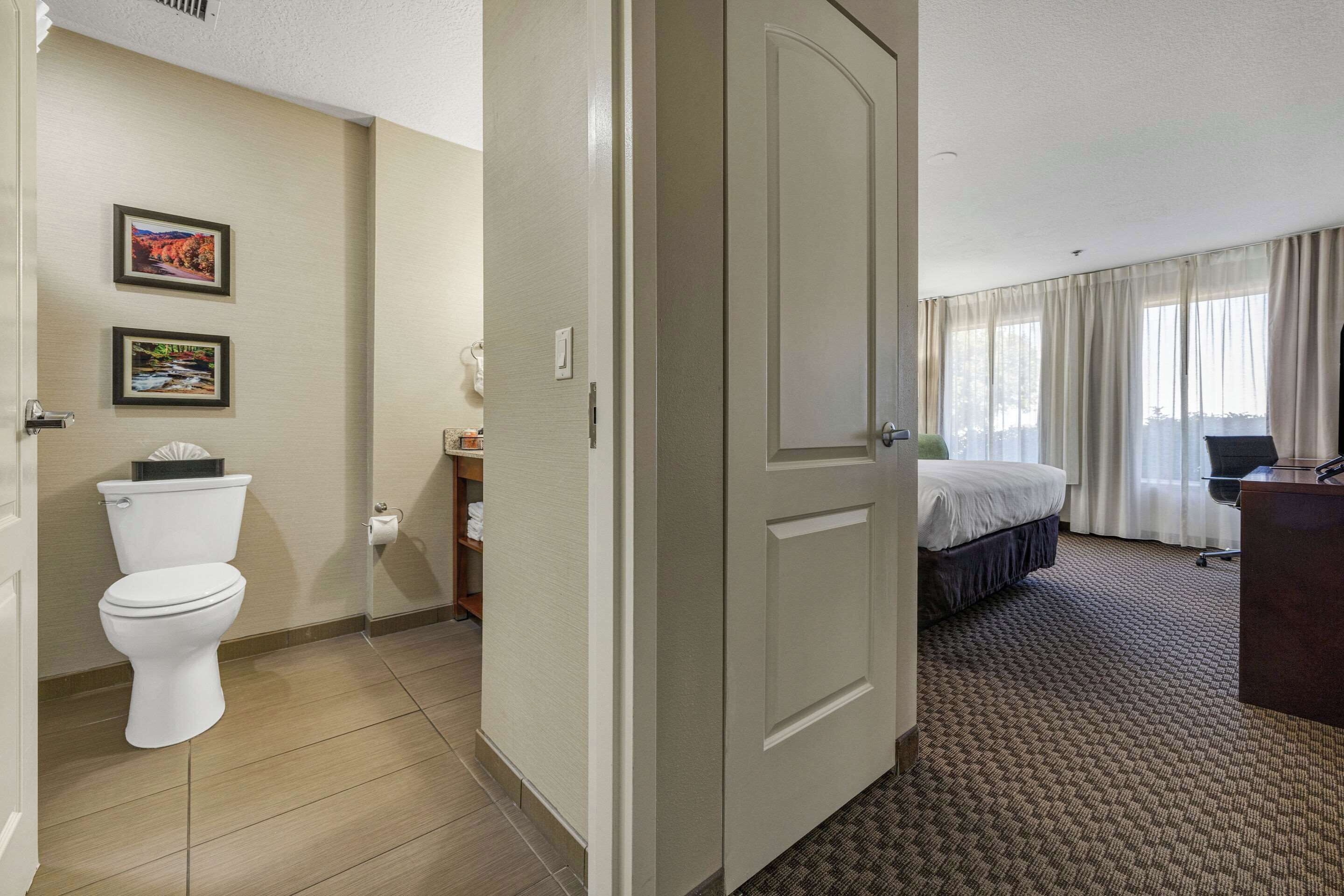 A hotel room with a toilet, a bed, a desk, and a window with curtains.