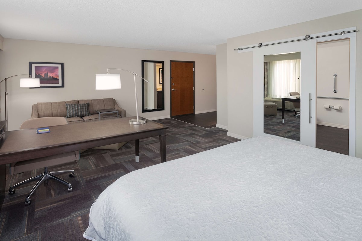 Une chambre d’hôtel avec un grand lit, un bureau avec une chaise, un canapé, un miroir et une porte donnant sur une autre pièce.