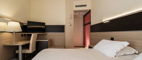 Una habitación de hotel con cama, escritorio, silla, televisión y un cubo de basura.