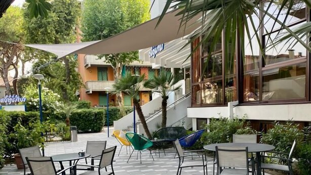 An outdoor patio area with tiled flooring, various seating options including wicker chairs and tables, and a building with a sign in a foreign language. - Hotel Augustus (Riccione)