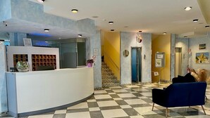 A reception area with a checkered floor, a reception desk, a blue chair, and a staircase in the background. - Hotel Augustus (Riccione)