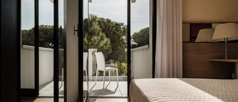 Habitación de hotel con cama, mesita de noche, lámpara y vista a un balcón con silla y árboles.