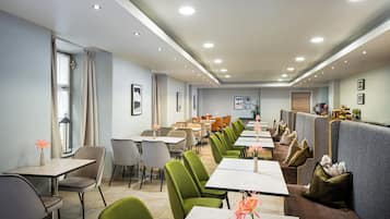 Uma área de refeições moderna com várias mesas e cadeiras, um balcão longo com um mostrador de menu e vista para a área de refeições através de uma janela.