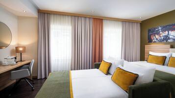 Et hotelværelse med to senge, et skrivebord, en stol, en lampe og et stort vindue med gardiner.