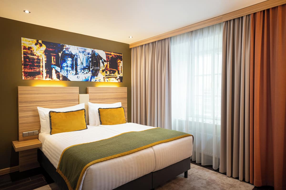 A modern hotel room with a large bed, a headboard with a cityscape artwork, a nightstand, and a view of the city through sheer curtains.