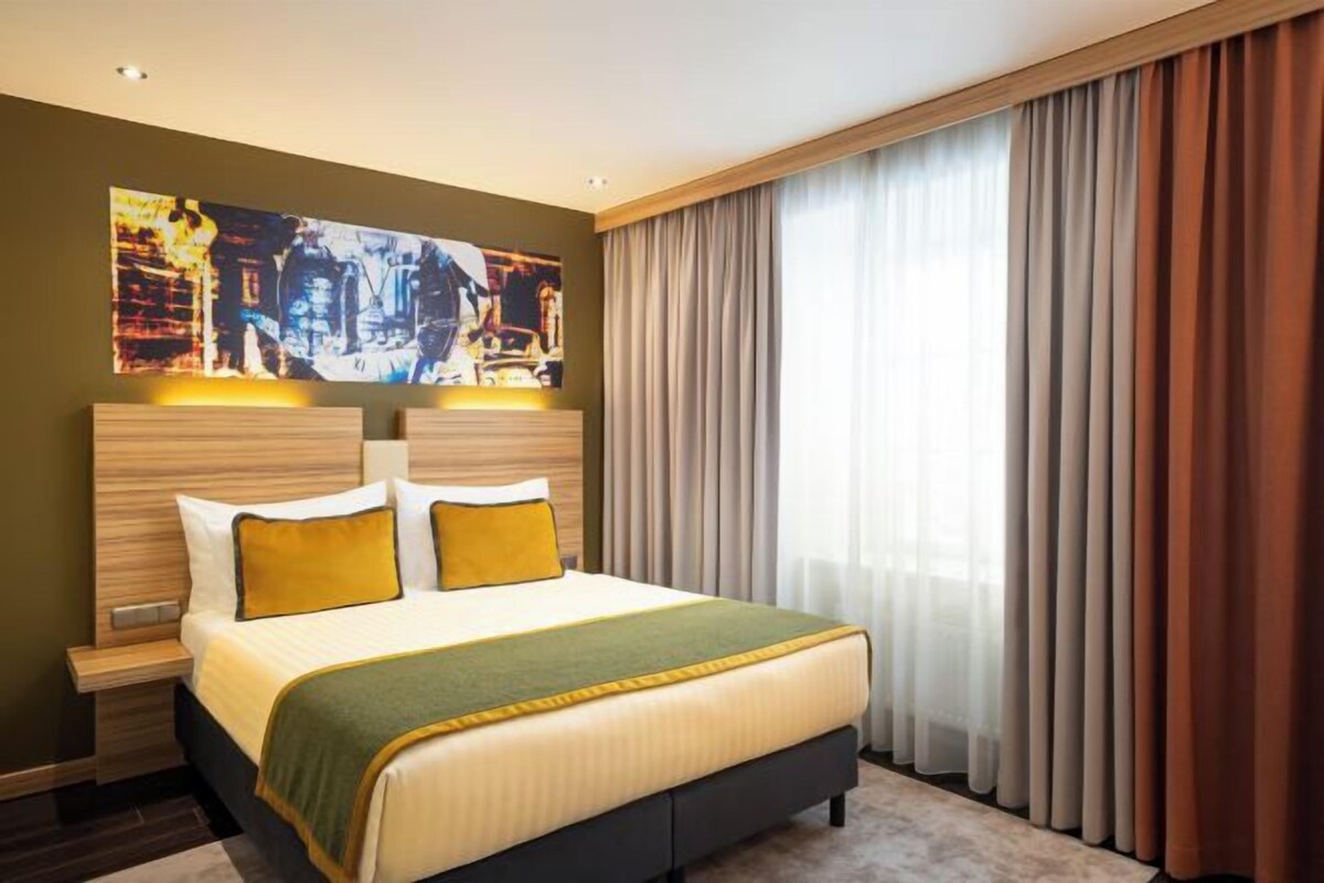 Habitación de hotel con una cama grande, una mesita de noche, un cuadro en la pared y una ventana con cortinas.