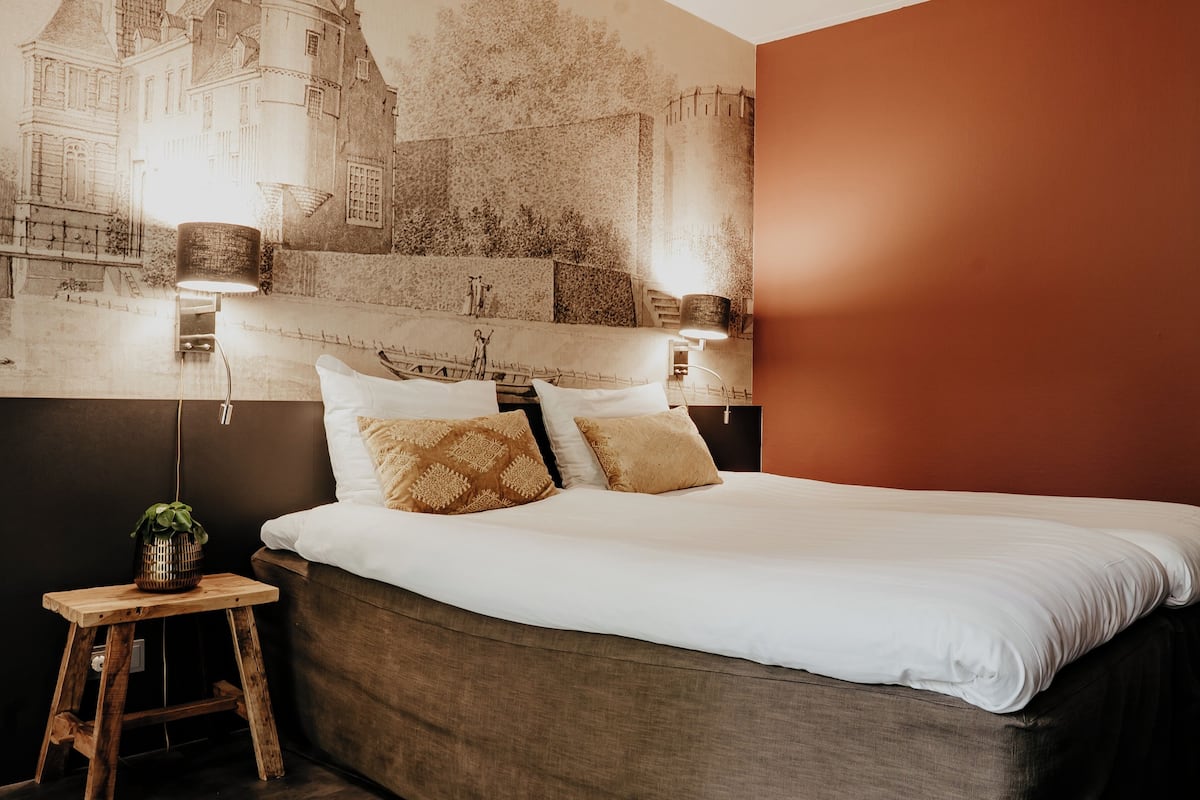 Ein Bett mit weißen Leinentüchern, zwei Zierkissen, ein Nachttisch aus Holz mit einer Pflanze und eine Wandlampe.
