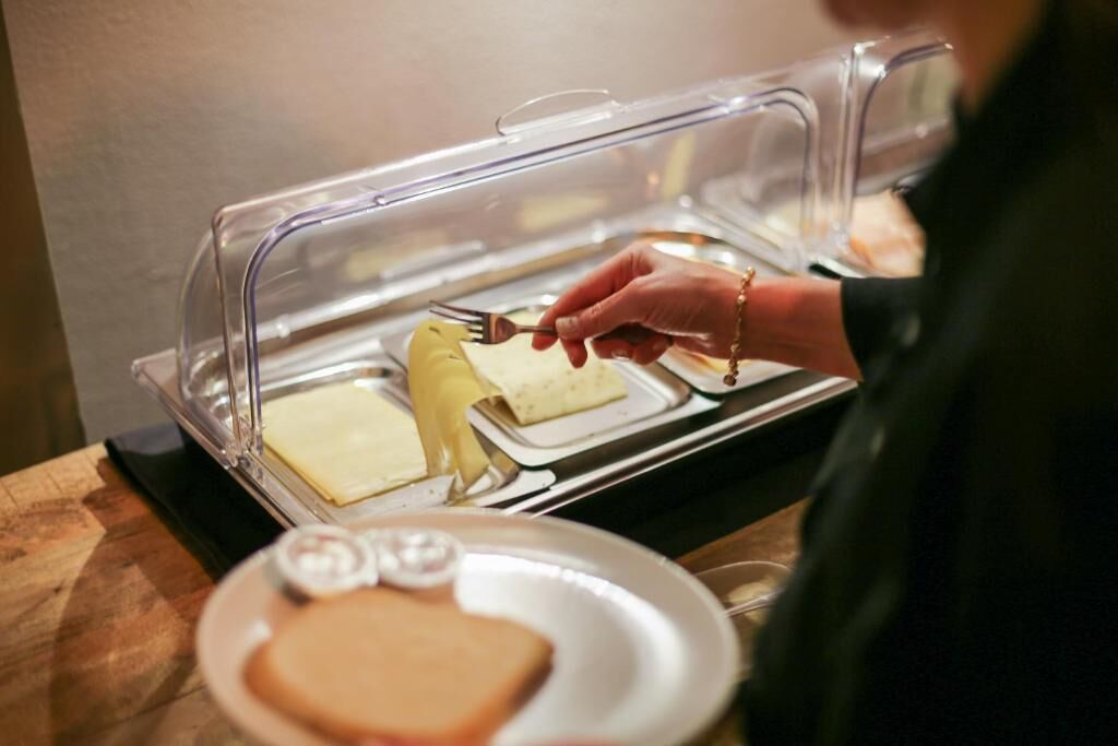 Ein Buffet mit einer transparenten Schutzabdeckung, ein Teller mit Brot und eine Hand, die eine Gabel hält.