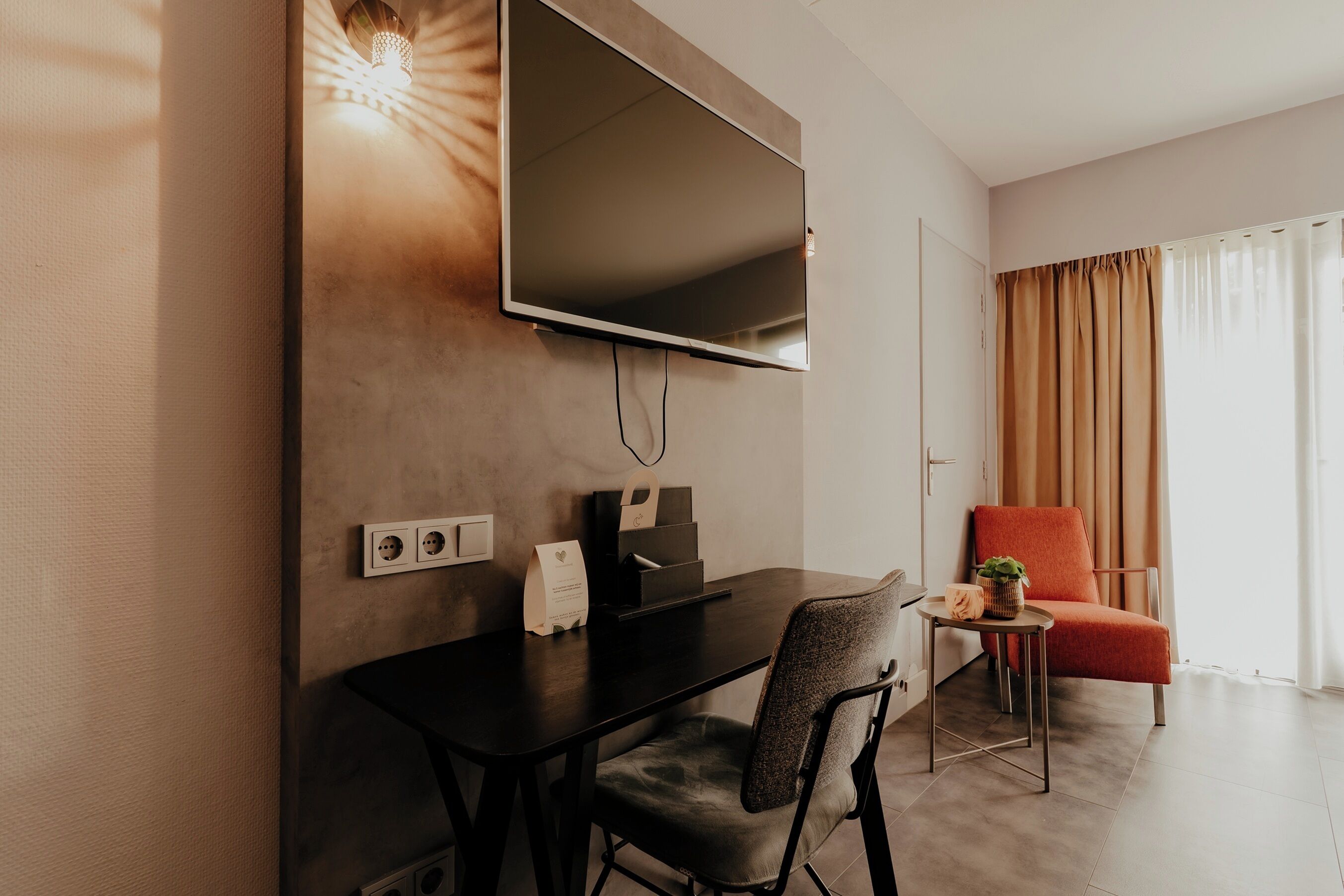 Una habitación moderna con un televisor de pantalla plana en la pared, un escritorio con una silla, una mesita con una planta y una silla roja.