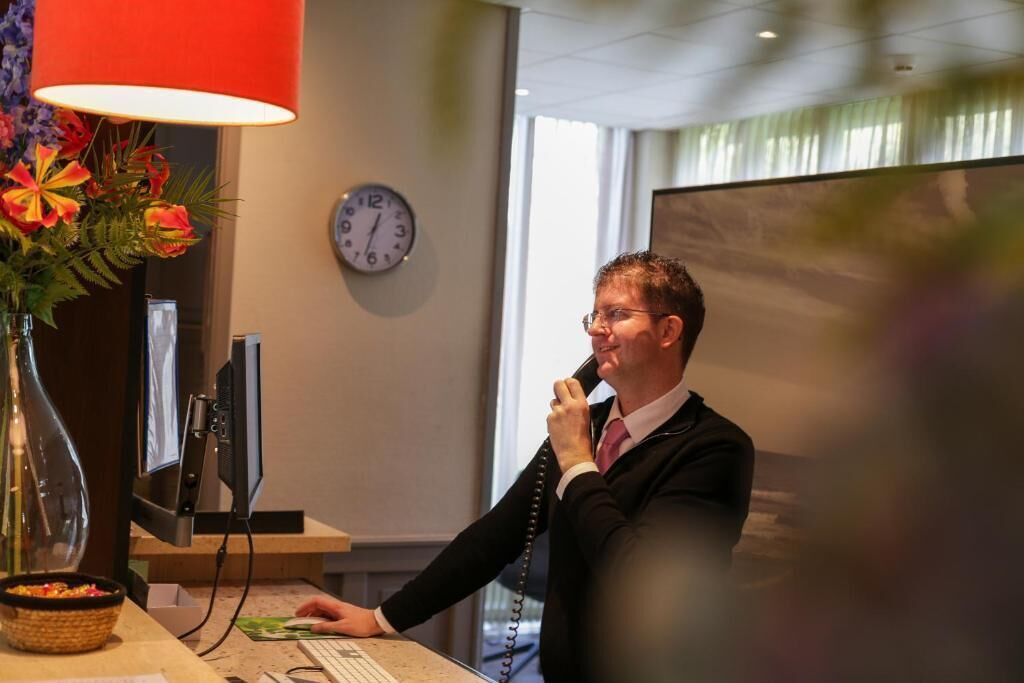 A man in a suit speaking on a telephone at a desk with a computer monitor, a clock, and a vase of flowers.