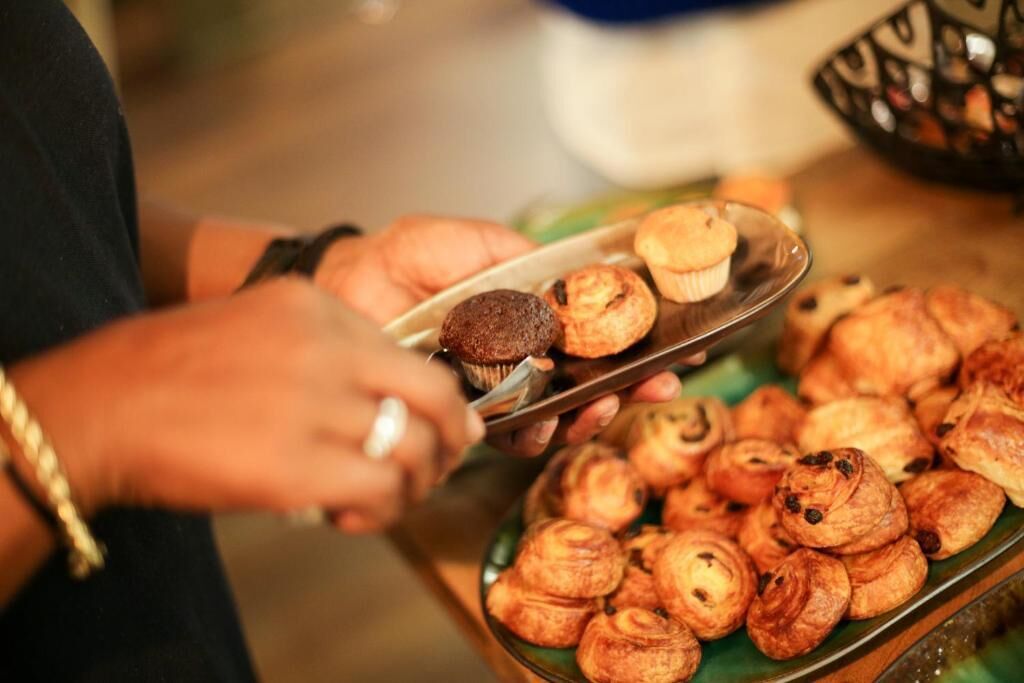 Eine Person hält ein Tablett mit Gebäck, darunter Muffins und Croissants, an einem Buffet.