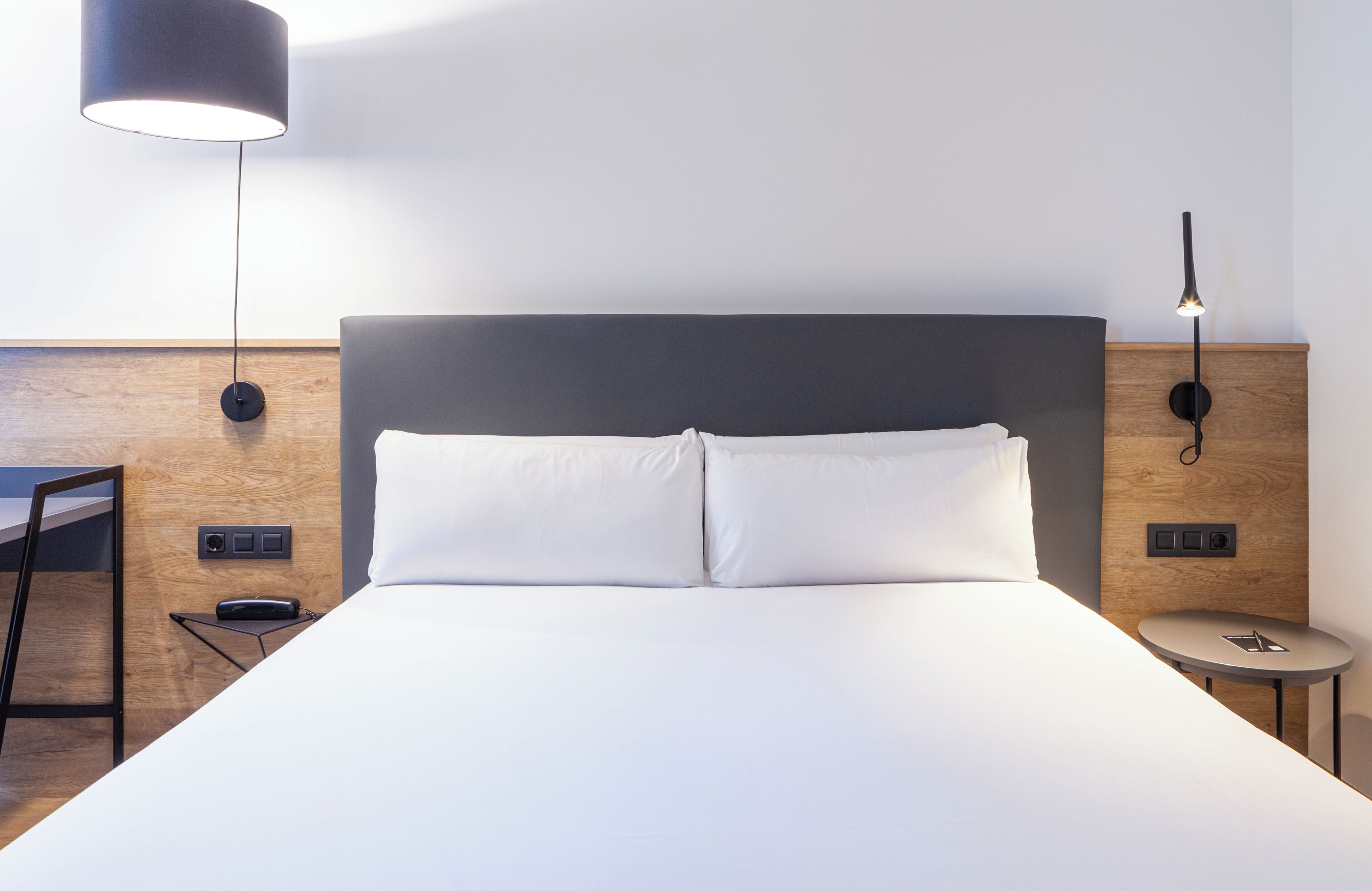A modern hotel room with a large bed, bedside tables, a desk, and a lamp.