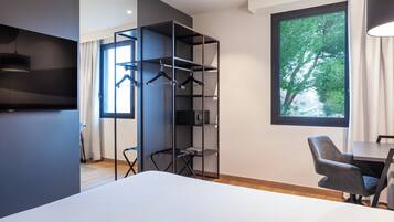 Una habitación de hotel moderna con cama, escritorio, silla, armario con ropa colgada y un amplio ventanal con vista a árboles.