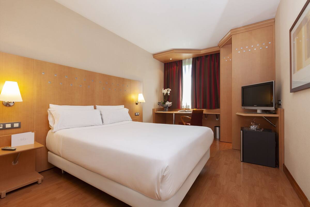 Ein Hotelzimmer mit einem großen Bett, Nachttischlampen, einem Schreibtisch mit Stuhl, einem Fernseher und einem kleinen Kühlschrank.