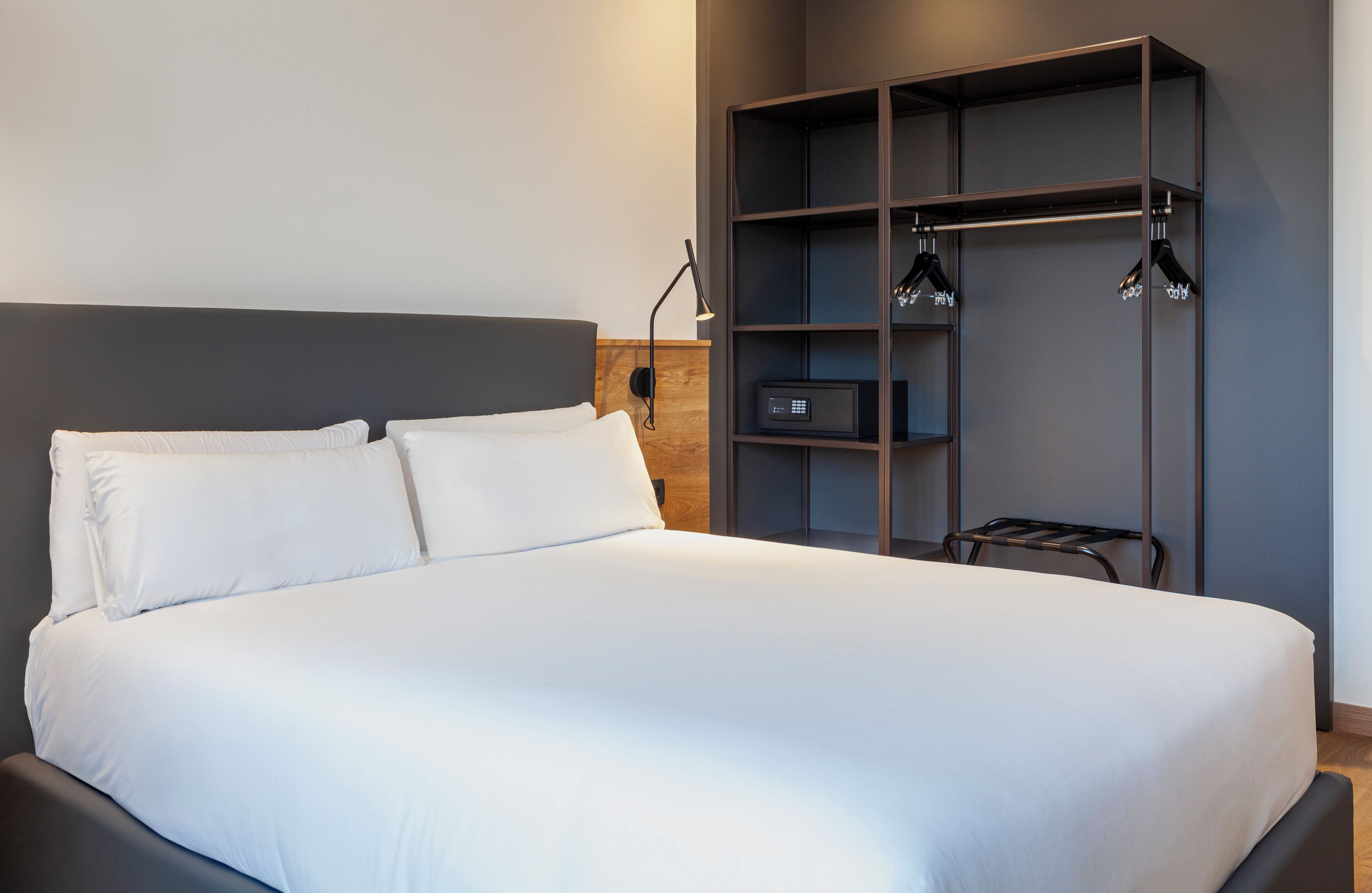 A modern hotel room with a large bed, a wooden headboard, a built-in shelf with a radio, and a wall-mounted lamp.
