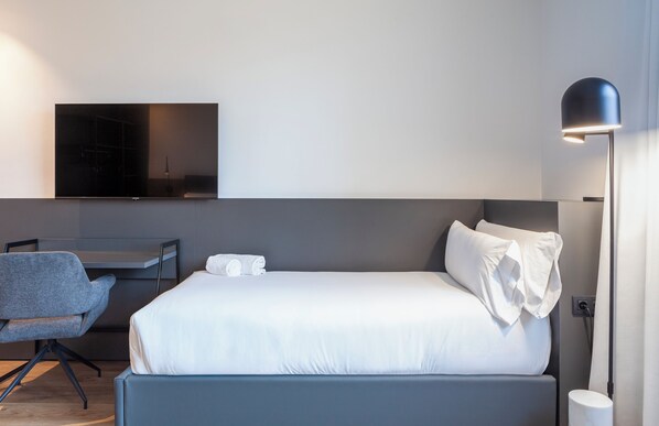 A modern hotel room with a bed, a desk, a chair, a flat-screen TV mounted on the wall, and a floor lamp.