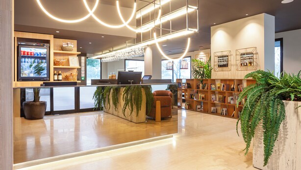 A modern reception area with a counter, seating, a refrigerator, shelves with items, and a large plant. - Hotel SB Madrid Norte (San Sebastian de los Reyes)