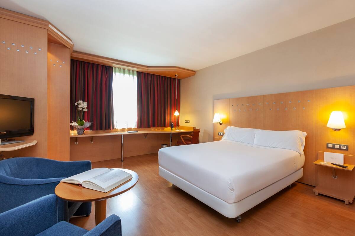 Ein Hotelzimmer mit einem Bett, einem Schreibtisch, einem Sessel, einem Fernseher und einem kleinen Tisch mit einem Buch.