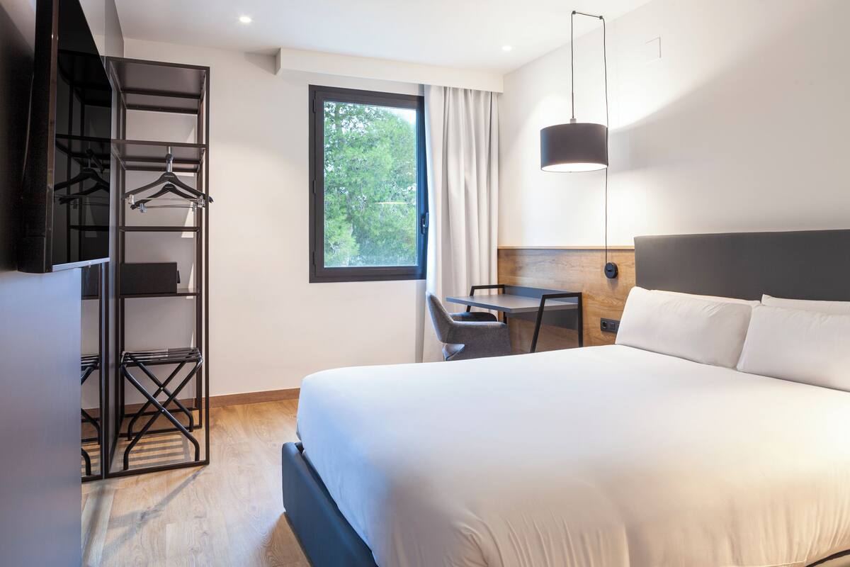 Ein modernes Hotelzimmer mit einem großen Bett, einem Schreibtisch, einem Kleiderschrank und einem Fenster mit Blick auf Bäume.