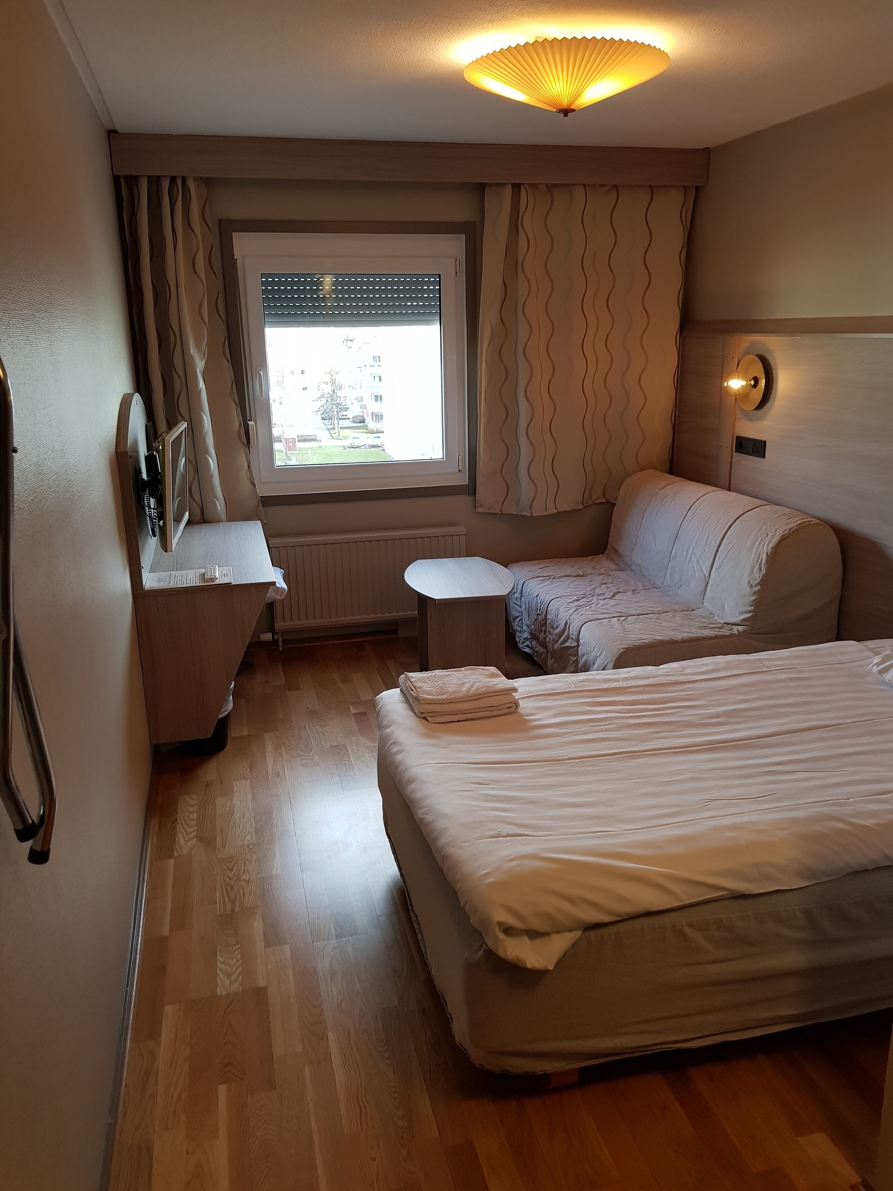 A hotel room with a bed, a desk, a chair, a small table, and a window with curtains.