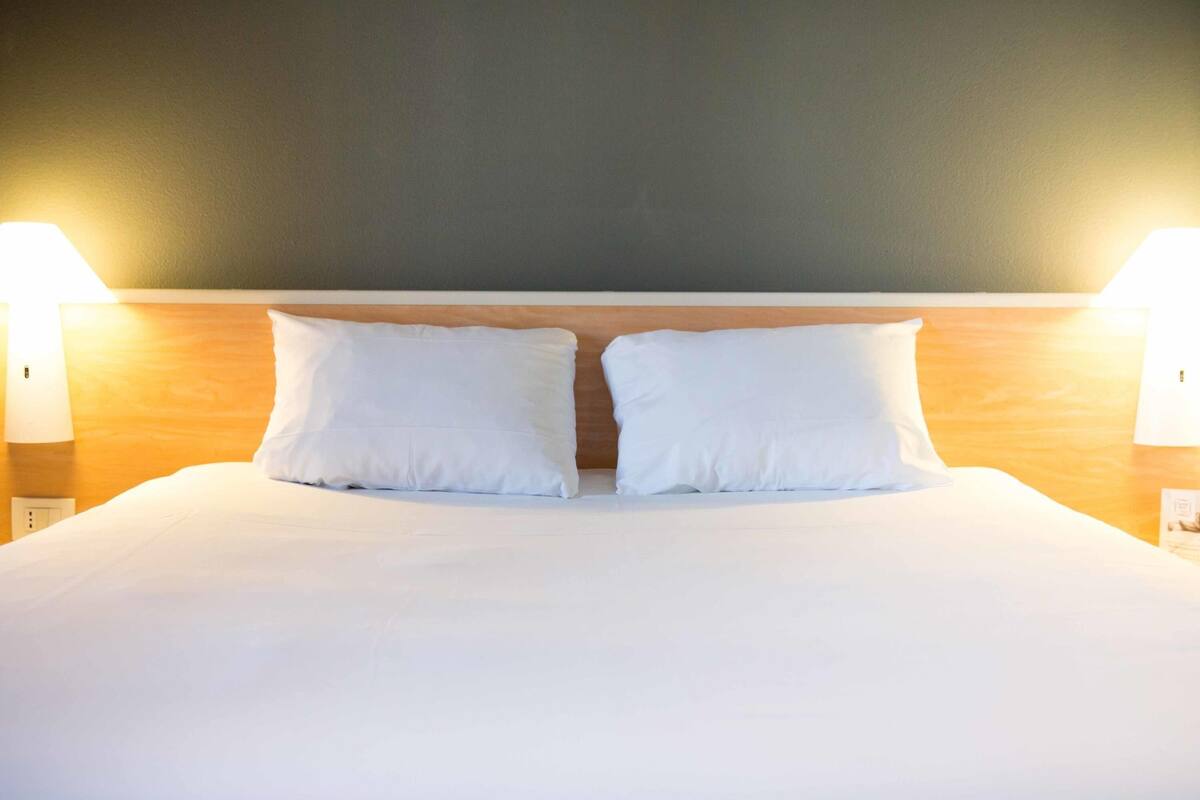 A neatly made bed with two pillows, a wooden headboard, and two wall-mounted lamps.