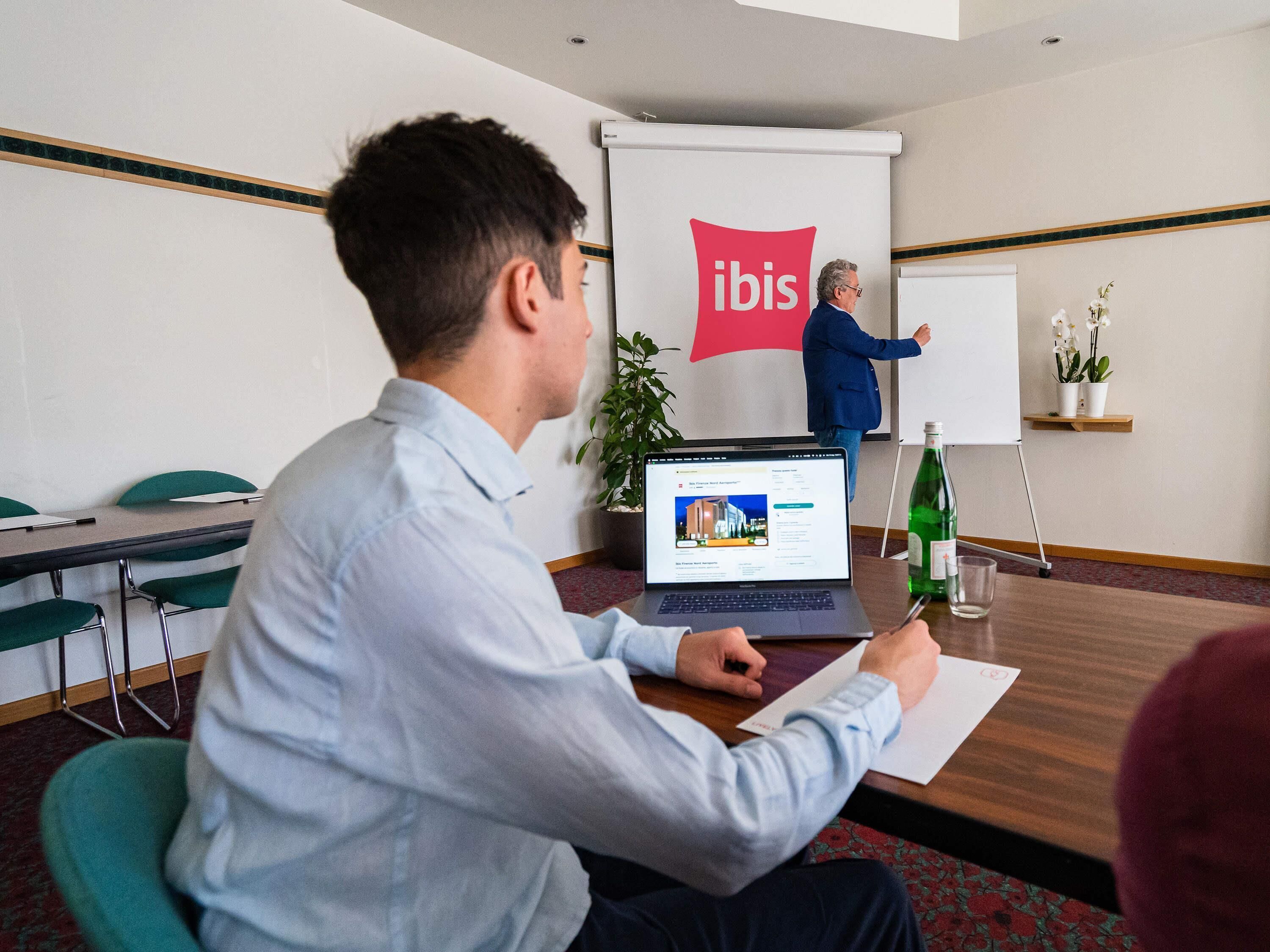 A conference room with a laptop, a whiteboard, and a presentation screen with the 'ibis' logo.