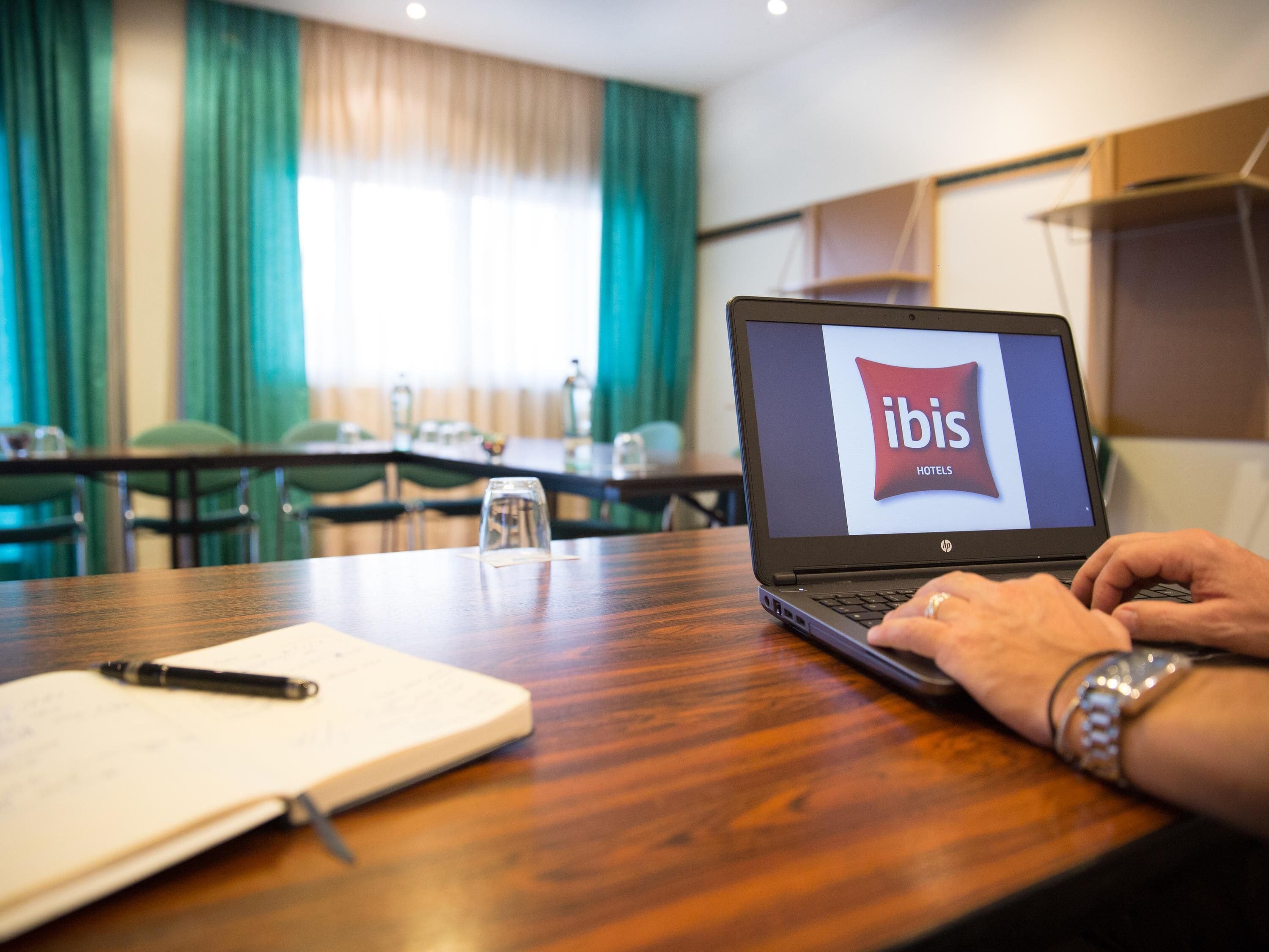 A laptop on a wooden table displaying the ibis hotels logo, a notebook, a pen, and a glass of water.