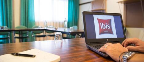 A laptop on a wooden table displaying the ibis hotels logo, a notebook, a pen, and a glass of water.