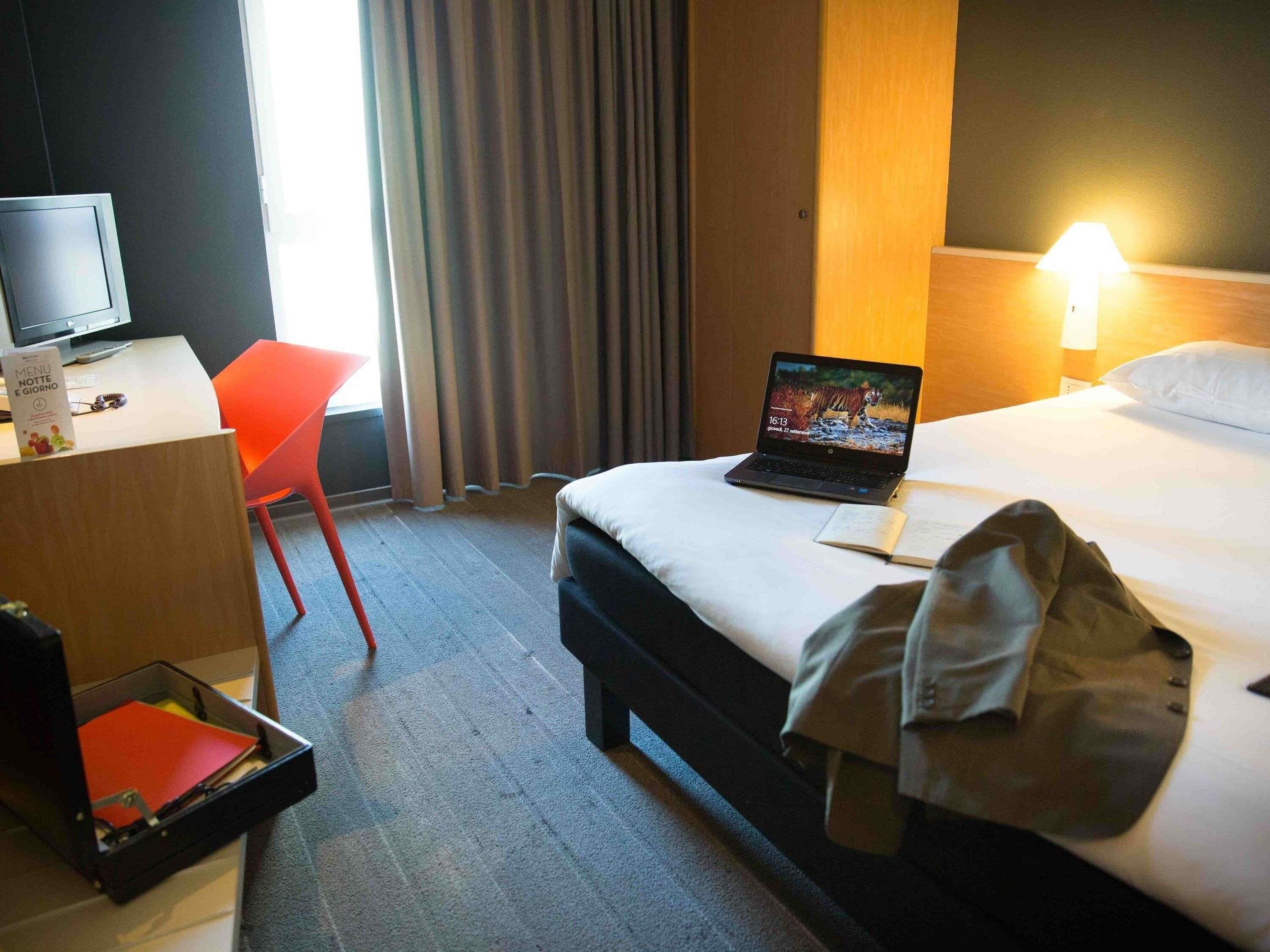 A hotel room with a bed, a desk with a laptop, a chair, and a bedside table with a lamp.