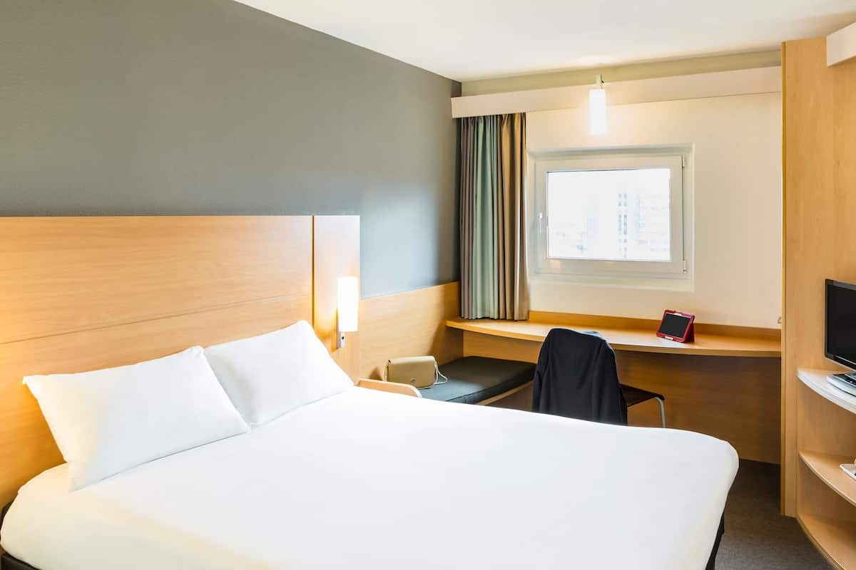 Habitación de hotel con una cama grande, un escritorio con silla, televisor y ventana con cortinas.