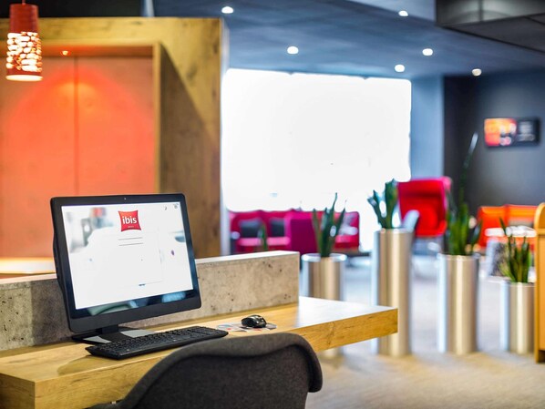 Un mostrador de recepción con computadora, teclado y ratón. Hay plantas en macetas y una silla roja al fondo.