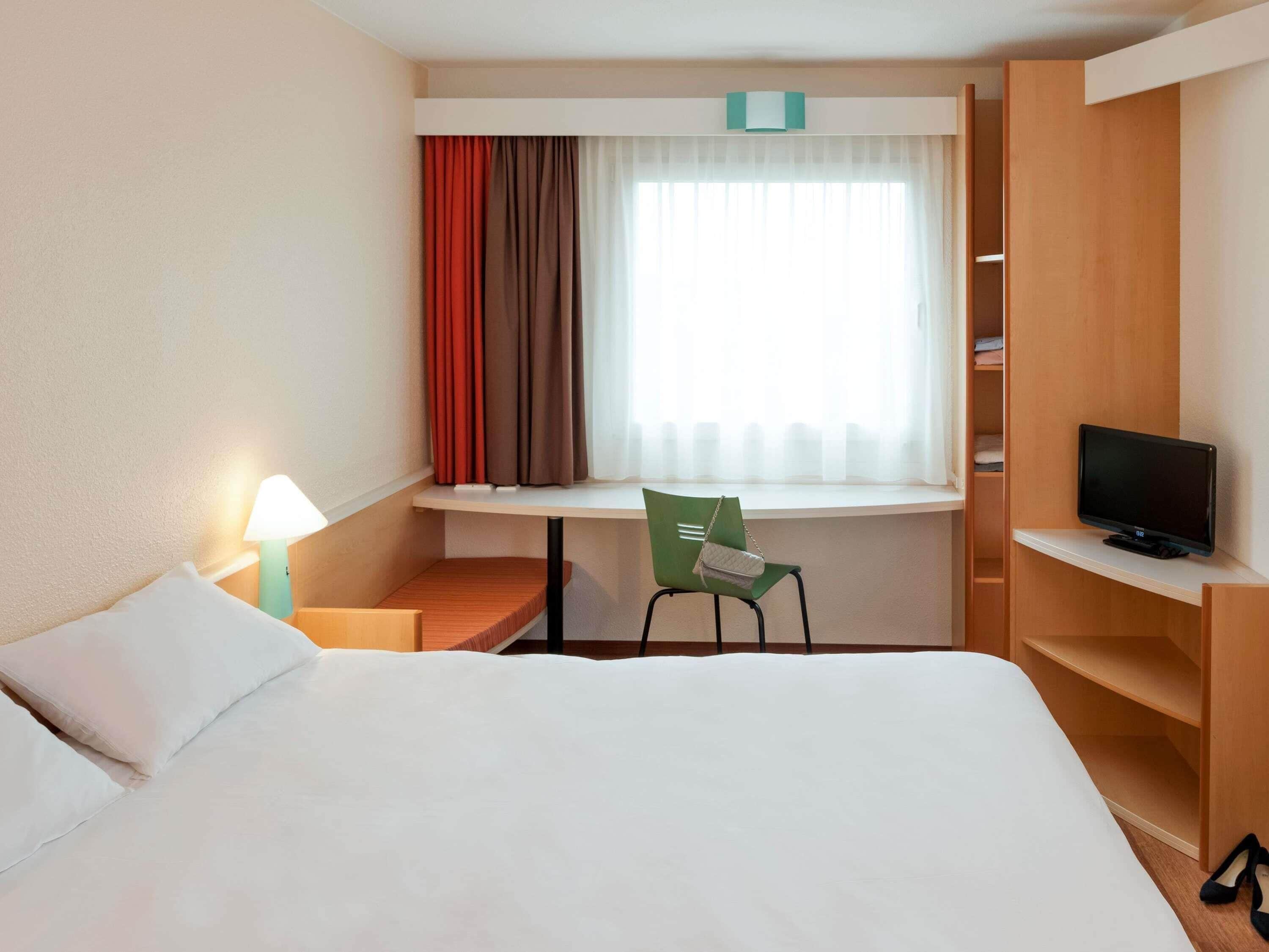 Une chambre d’hôtel avec un lit, un bureau, une chaise, une télévision et une fenêtre avec des rideaux.
