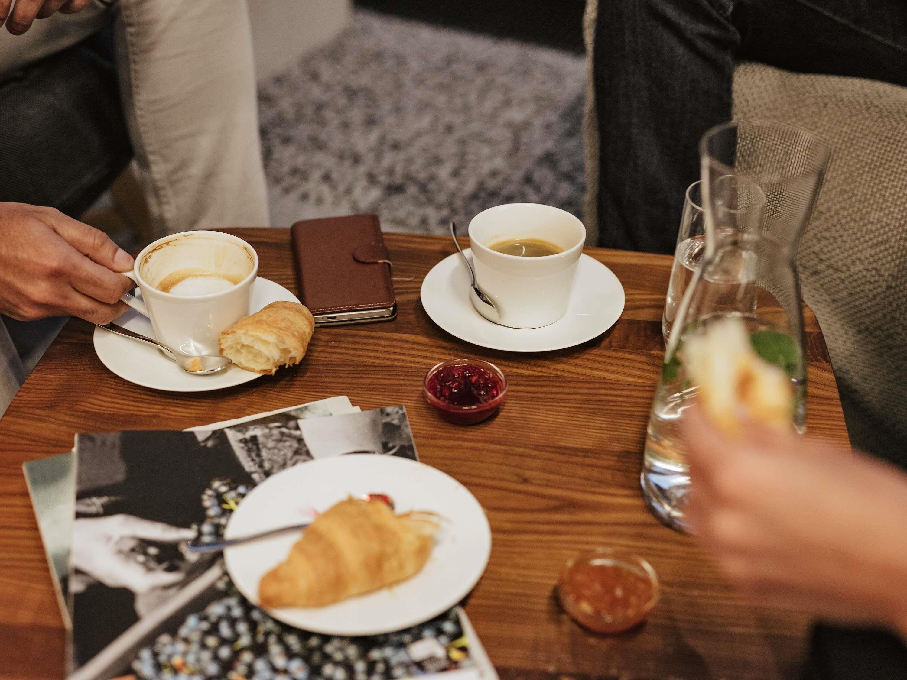 Una mesa de madera con una taza blanca, una cartera marrón, un croissant, un vaso de agua y un recipiente pequeño de mermelada.